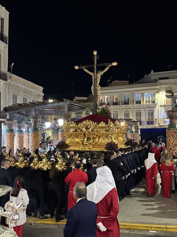 El trono del Cristo de la Paz avanzando por la plaza con los portadores debajo y los nazarenos de túnica roja a los lados
