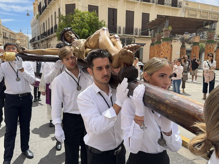 Jóvenes de camisa blanca llevando en brazos la cruz del Cristo de la Paz por una calle de Melilla