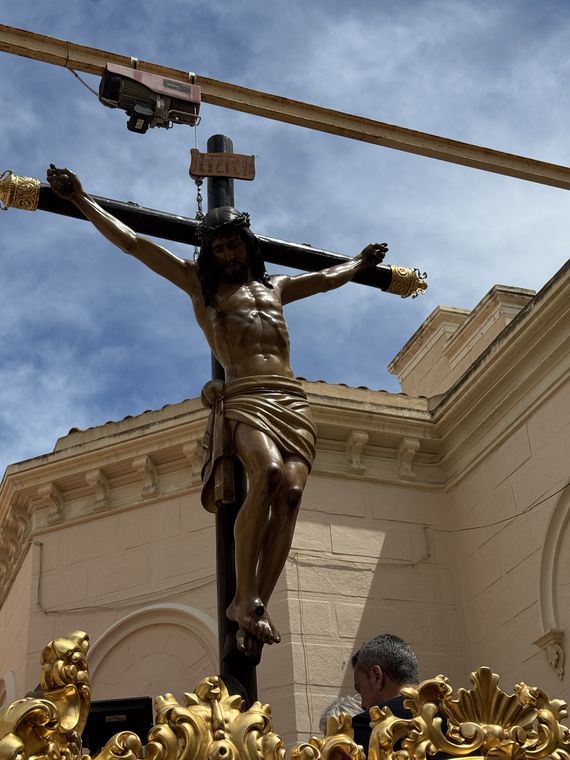 El Cristo de la Paz sobre su cruz ante la fachada de la parroquia, bajo un cielo con nubes