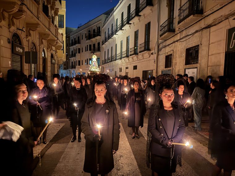 Mujeres de mantilla con velas encendidas caminando por una calle de Melilla, con el trono al fondo