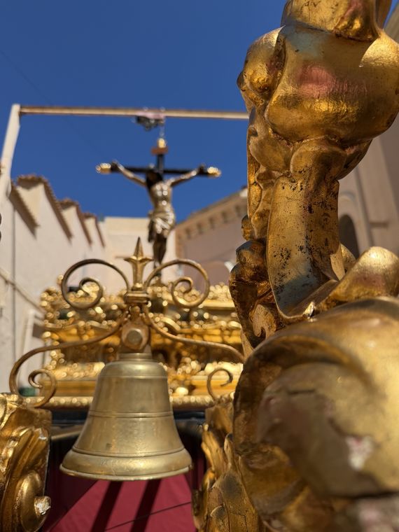 Detalle dorado del trono con su campana y, al fondo, el Cristo crucificado contra un cielo azul